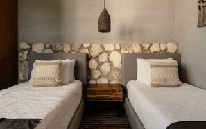 Ein Zimmer mit zwei Betten, einer Steinsäule, einer gewebten Pendelleuchte und einem kleinen Holztisch.