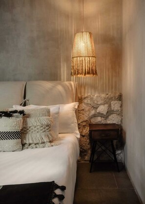 Een slaapkamer met een bed, kussens, een hanglamp, een stenen muur en een klein houten tafeltje.