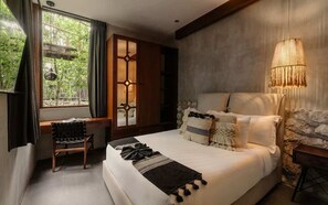 A bedroom with a bed, a desk, a chair, a window with curtains, a wall-mounted lamp, and a stone accent wall. - Siwa Jungle Rain A2-004 Beach Club Access (Tulum)