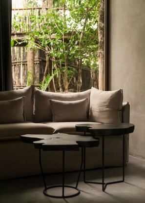 A sofa with multiple cushions, a small round table, and a window with a view of greenery. - Siwa Jungle Rain A2-004 Beach Club Access (Tulum)