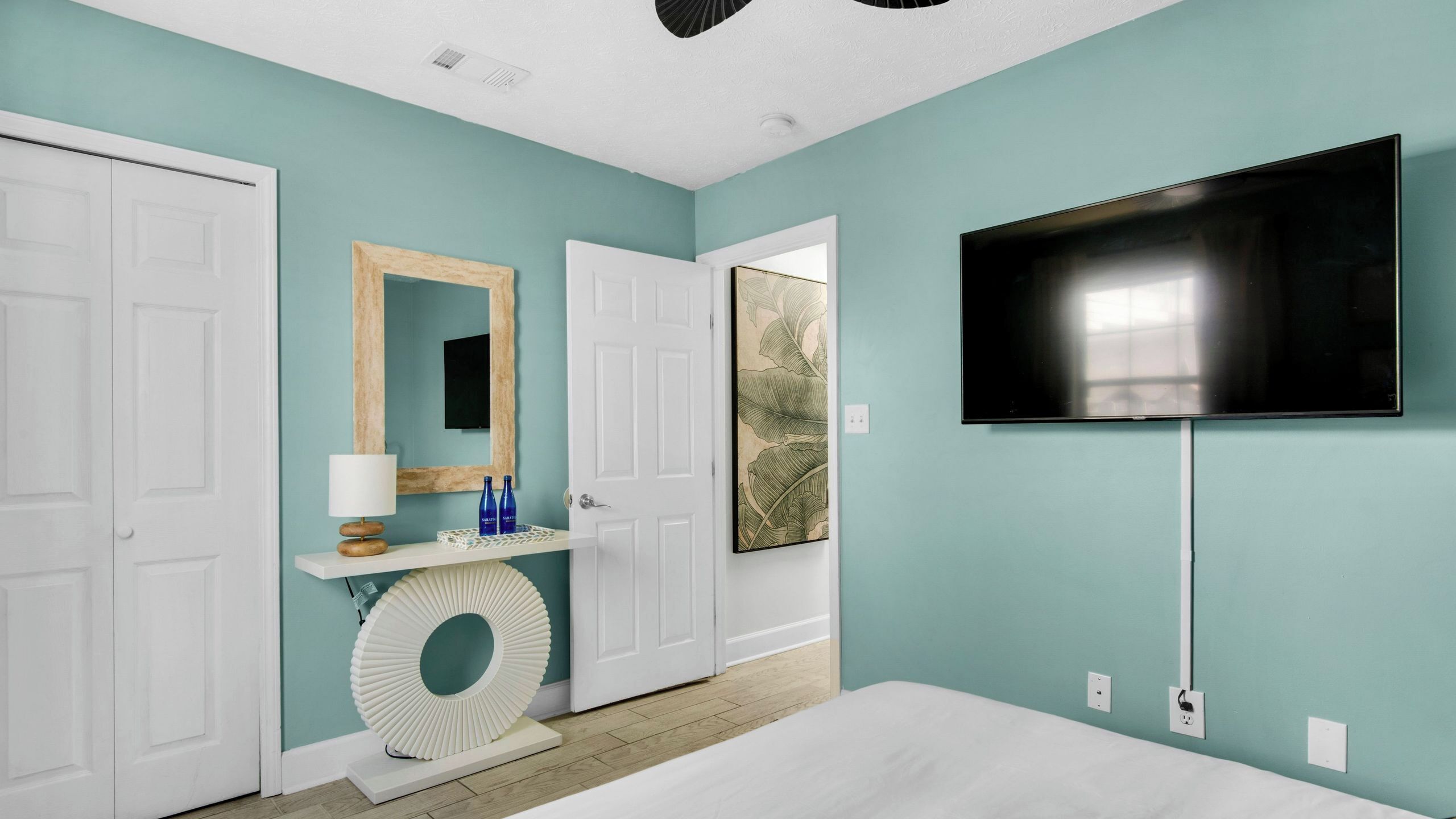 A bedroom with a bed, a TV, a door, a mirror, and a wall with a picture.