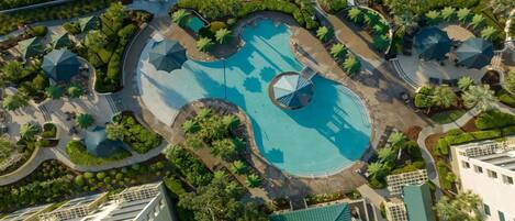 An aerial view of a resort with multiple swimming pools, surrounded by lush greenery and shaded by umbrellas.