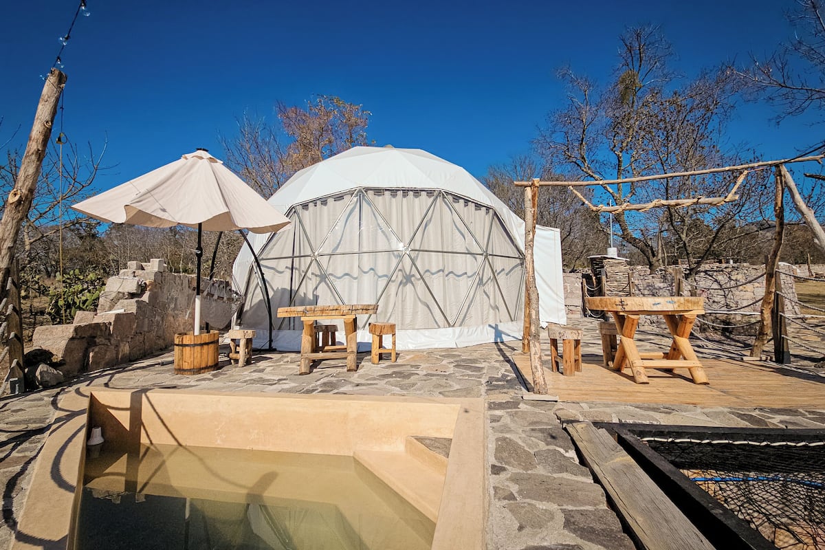 Una estructura de domo geodésico con piscina, mesas de madera y suelo de piedra.