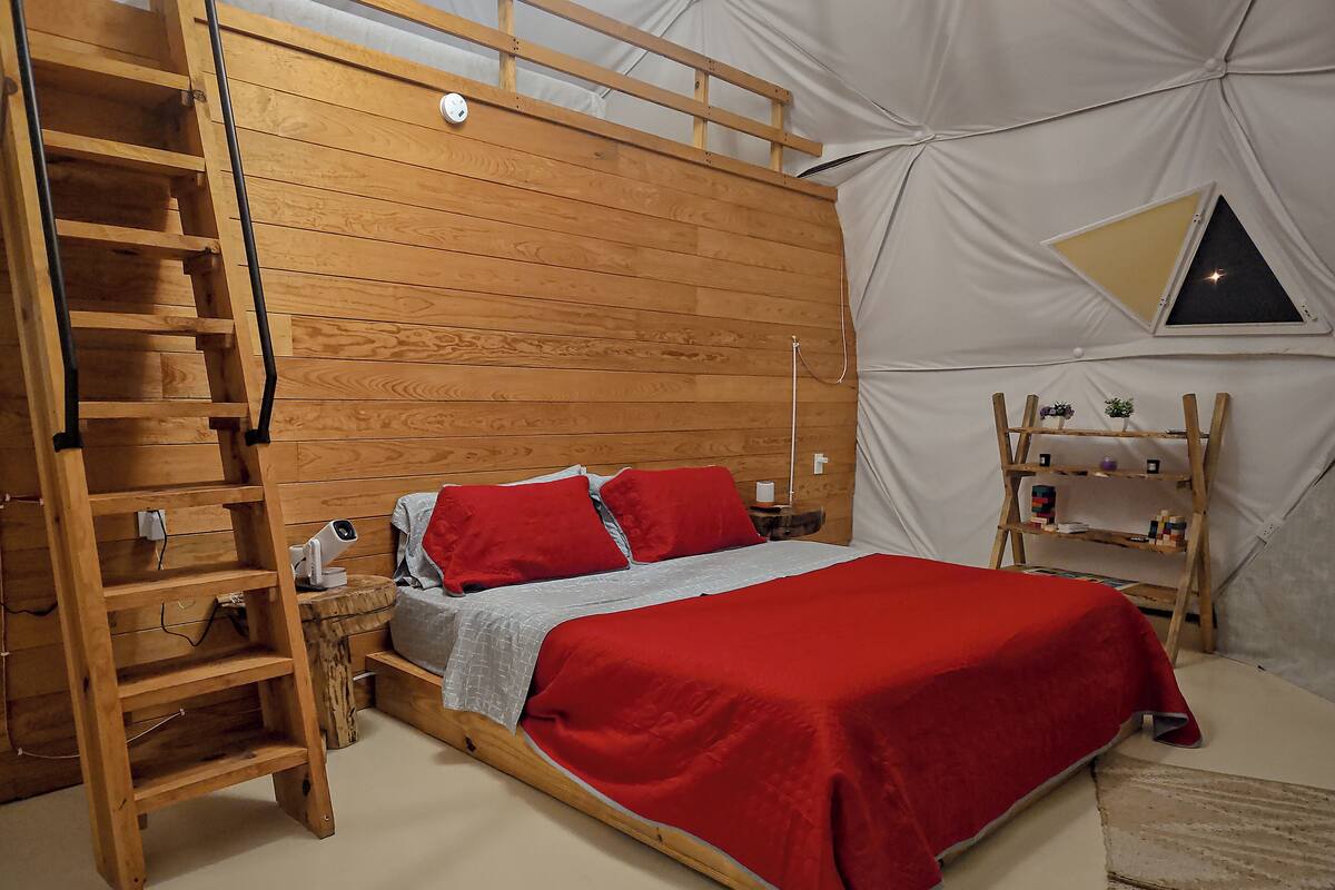 Una habitación con una cama de madera, una escalera, un estante pequeño y una ventana triangular.
