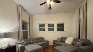 Ein Wohnzimmer mit zwei grauen Sofas, Holzboden, Deckenventilator und einem Fenster mit Vorhängen.