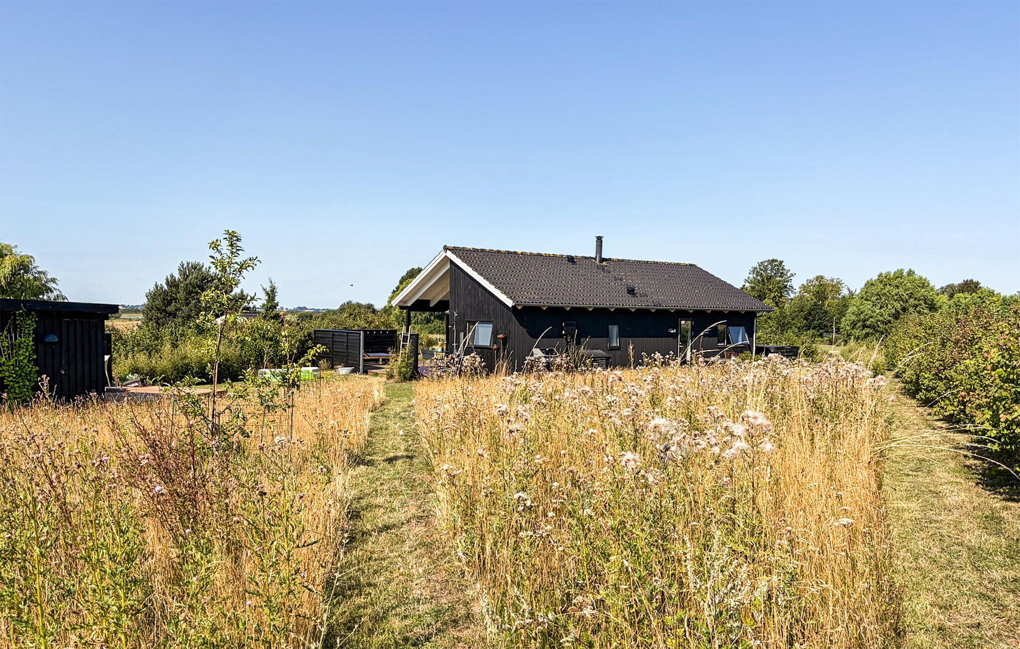Una casa rural amb teulada fosca i un petit cobert annex, envoltada d'herbes altes i arbres.