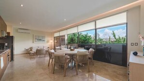 A spacious living area with a large dining table surrounded by chairs, a kitchen area, and a sliding glass door leading to a balcony with a view of greenery. - Near Tulum Beach 2BR –Private Pool & Cenote Access (Tulum)