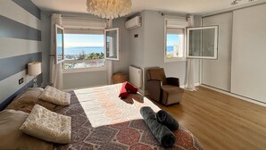 Ein Schlafzimmer mit einem großen Bett, einem Sessel, Meerblick und einem Fenster mit Vorhang.