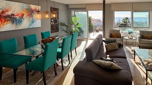 Ein modernes Wohnzimmer mit einem Glastisch, smaragdgrünen Sesseln, einem grauen Sofa und einem großen Fenster mit Meerblick.