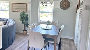 Een eetruimte met een houten tafel, witte stoelen, een kroonluchter, een potplant en een klok aan de muur.