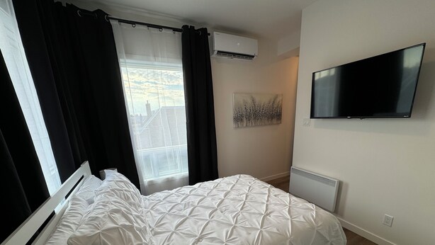 Ein Schlafzimmer mit Bett, schwarzen Vorhängen, einem Fenster, einer Klimaanlage, einem Wand-Fernseher und einem Heizkörper.