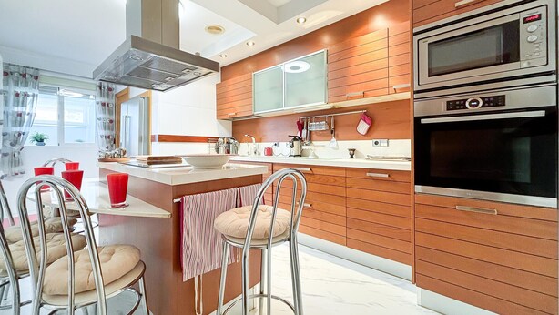 Une cuisine moderne avec des meubles en bois, des appareils en acier inoxydable, un îlot central avec des tabourets et une fenêtre avec des rideaux.
