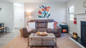A living room with a brown leather sofa, a coffee table with decorative items, a fireplace, and a wall-mounted painting. - Ballantyne Retreat – Updated 2BR/2BA Near Shops & Medical (Charlotte)