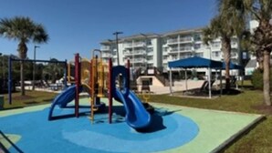 Ein Spielplatz mit Rutsche und Klettergerüsten, umgeben von Palmen und einem Gebäude im Hintergrund.