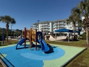 A playground with a slide and climbing structures, surrounded by palm trees and a building in the background. - Beachfront Condo w/ Balcony & Pool Access (Pawleys Island)