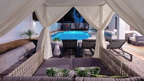 Uma área de estar coberta ao ar livre, com piscina, cortinas brancas e espreguiçadeiras.