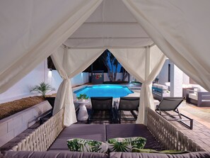 Un espace extérieur aménagé, abrité et doté d’une piscine, de rideaux blancs et de chaises longues.