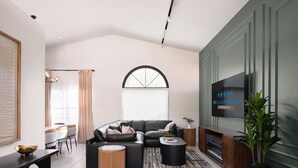 Uma sala de estar moderna com um sofá seccional, uma mesa de centro redonda de madeira, uma mesinha lateral preta e uma TV fixada em uma parede verde.
