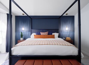 Ein Bett mit Baldachin, dessen weißes und orangefarbenes Karomuster von zwei Nachttischen mit Lampen eingerahmt wird, in einem Zimmer mit dunkelblauem Holzvertäfelung und einem großen Fenster mit Vorhängen.