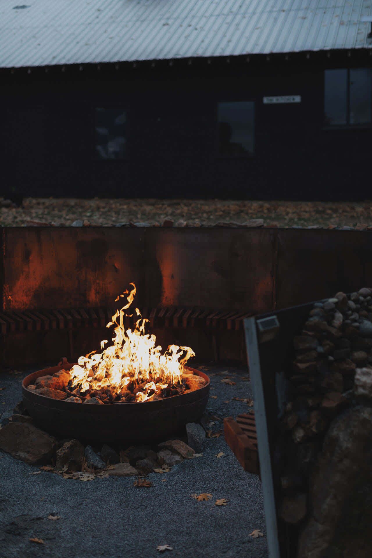 A fire pit with flames, surrounded by rocks and gravel, in an outdoor setting with a building in the background.