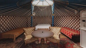 L’intérieur d’une yourte traditionnelle est aménagé avec une table ronde, deux fauteuils rembourrés, un lit recouvert d’une couverture à motifs rouges et un mur en treillis.