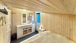 Una habitación con paneles de madera, una cama, un aparador con cestas, un espejo y una ventana con cortinas azules.