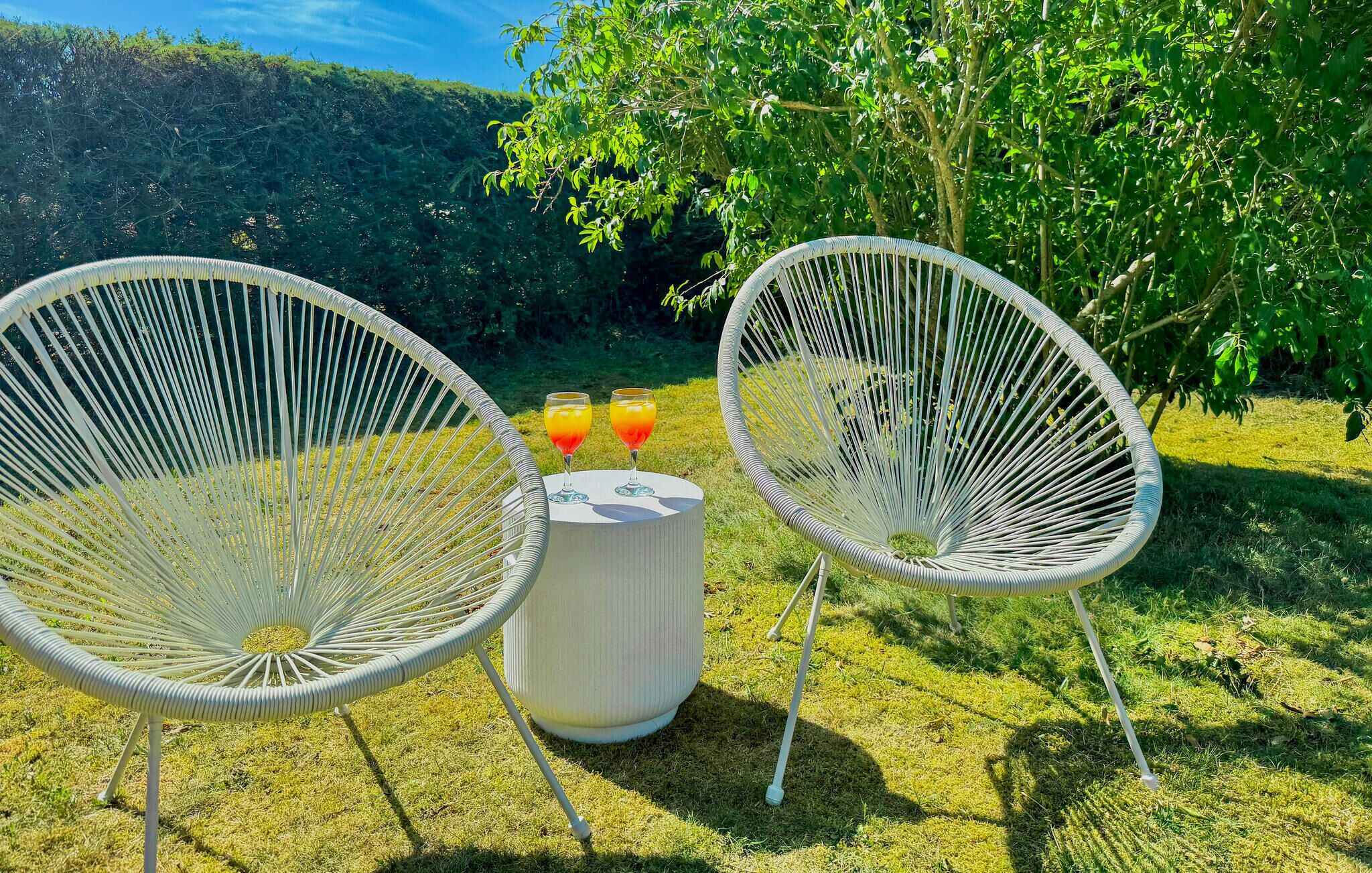 Zwei weiße Korbstühle mit einem Tisch und einem weißen zylindrischen Gegenstand dazwischen, auf einer grünen Fläche mit Bäumen im Hintergrund.