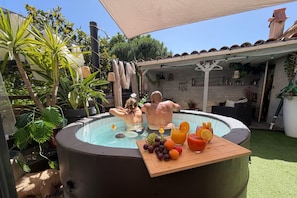 Et udendørs spa med et træbord med frugt og drikkevarer, omgivet af grønt og en overdækket terrasse.
