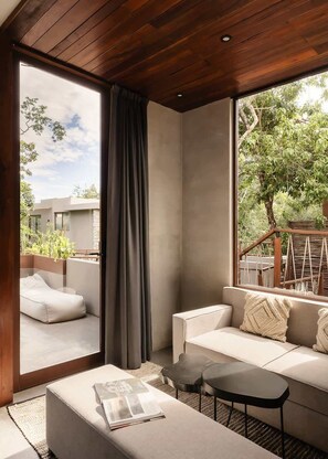 Ein modernes Zimmer mit Holzboden, einem großen Fenster mit Blick ins Grüne, einer Couch, einem kleinen Tisch und einer Leseecke.