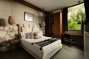 A bedroom with a stone wall, a bed with white bedding, a wooden headboard, a desk, and a view of greenery outside. - Siwa Jungle Paradise Retreat C1-003 Beach Club (Tulum)