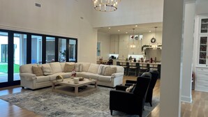 A spacious living room with a large sectional sofa, a coffee table, and a black armchair. The room features a high ceiling with chandeliers, large windows, and a view of the outdoors. - SPACIOUS 4-bdrm sleeps 10+,heated porch & tv’s, pool,hot tub,play room (Oxford)