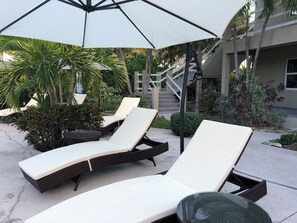 A sunlit outdoor seating area with lounge chairs and a table under a large umbrella, surrounded by greenery and a staircase. - ROSE COTTAGE-Tranquility steps from Grace Bay Beach and Grace Bay Strip! (Grace Bay)