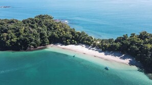 An aerial view of a tropical beach with clear turquoise waters, a sandy shore, and dense greenery.
