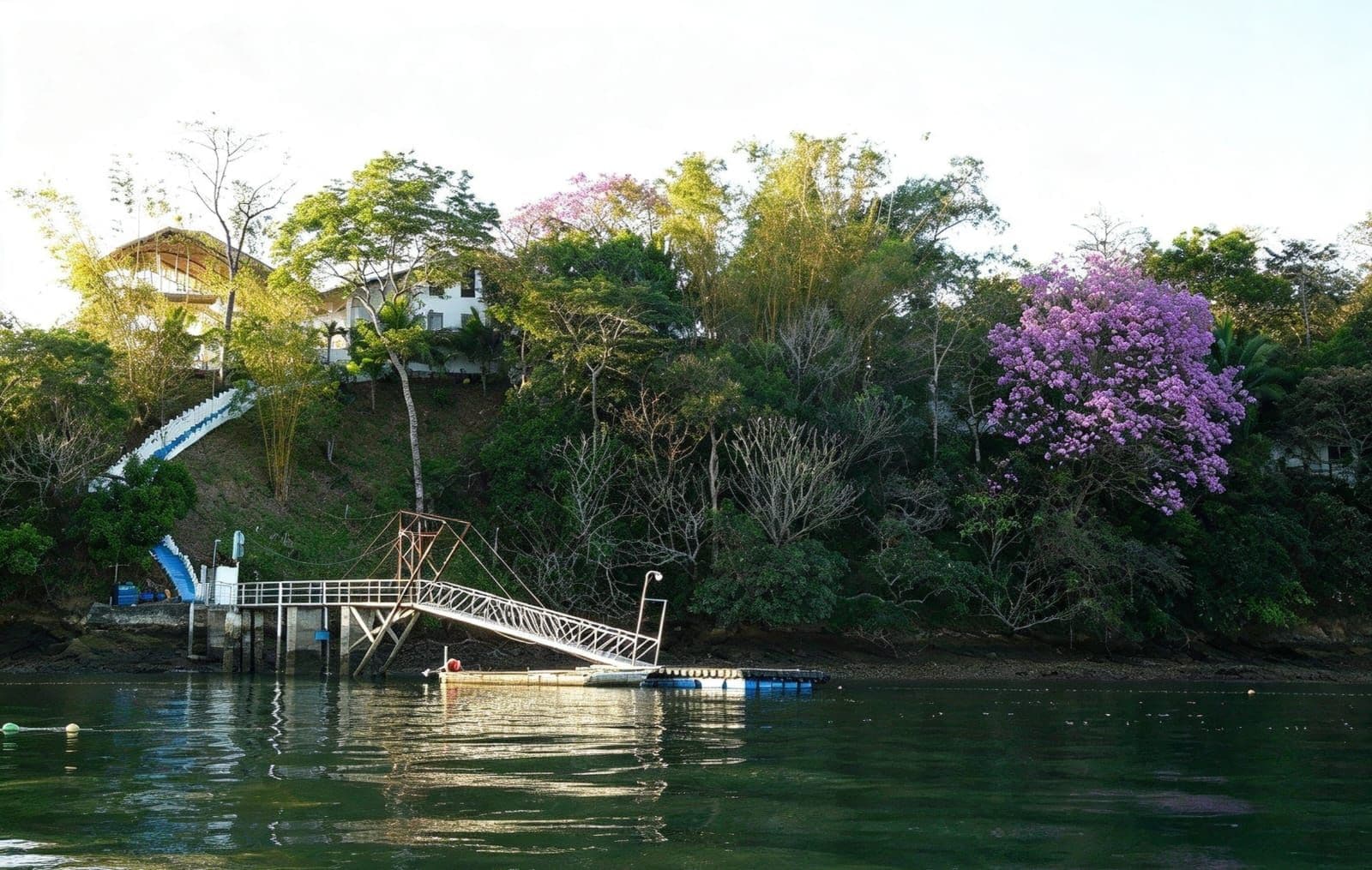 Un pont enjambant une étendue d’eau, avec un quai, entouré d’arbres et un bâtiment visible au loin.