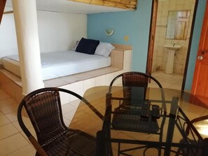A room with a bed, a glass table with chairs, a small sofa, and a bathroom with a sink.