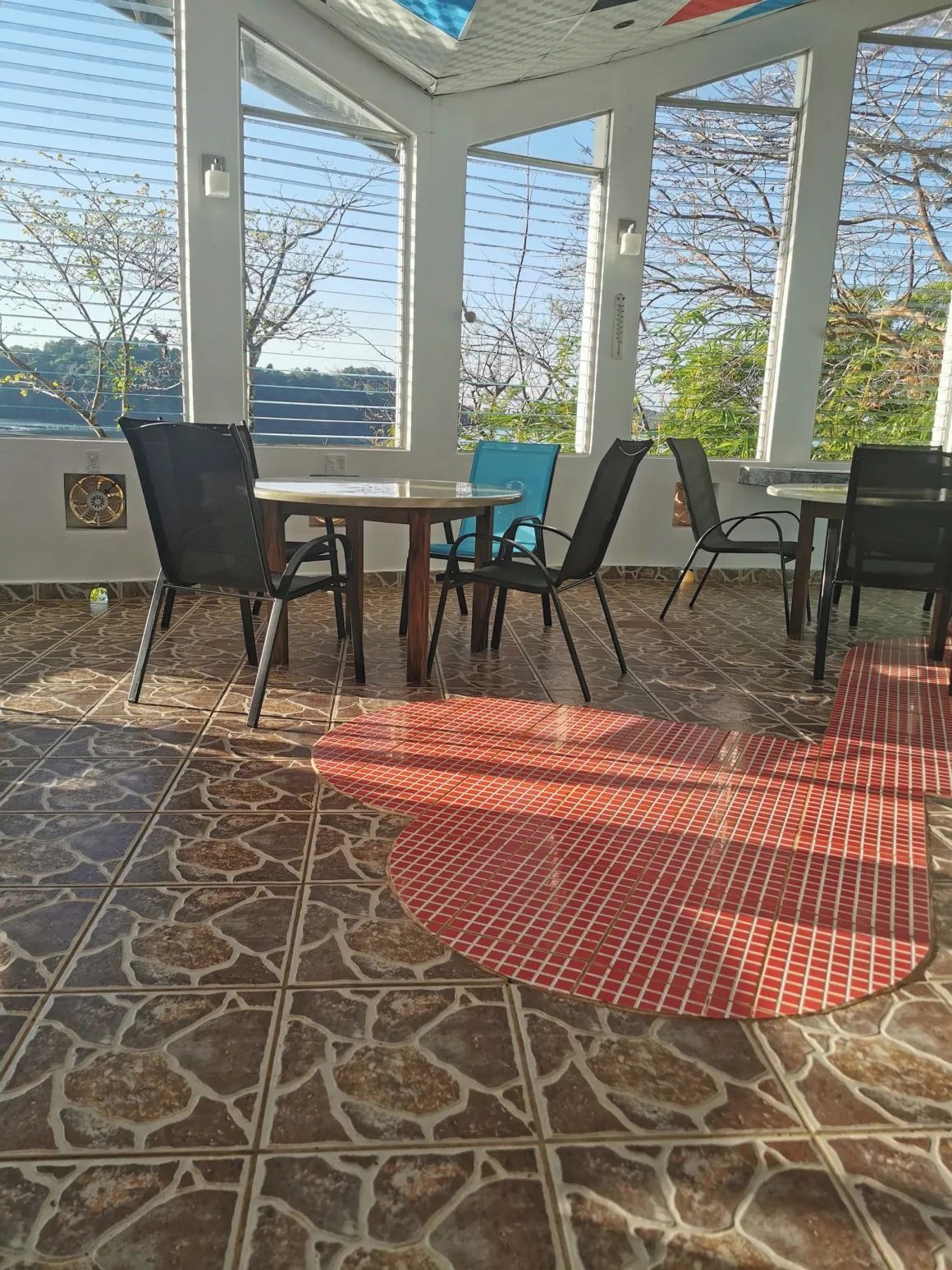Uma área de varanda coberta com mesa, cadeiras, janelas amplas e vista para o exterior.
