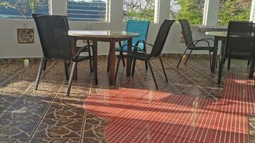 Uma área de varanda coberta com mesa, cadeiras, janelas amplas e vista para o exterior.