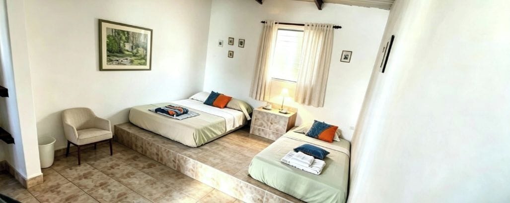 A bedroom with two beds, a chair, a window with curtains, and a painting on the wall.