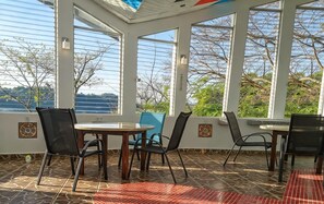 A sunlit patio with a view of the sea, featuring a round table and chairs, and a tiled floor.