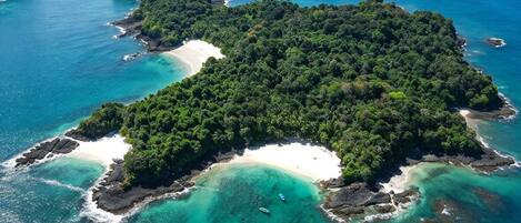 An aerial view of a tropical island with a central white sandy beach surrounded by dense green vegetation and clear turquoise waters.