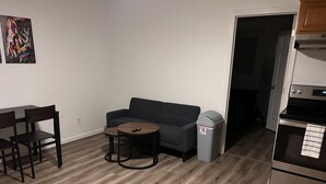 A compact living space with a sofa, a small round table, a trash bin, and a kitchenette area.