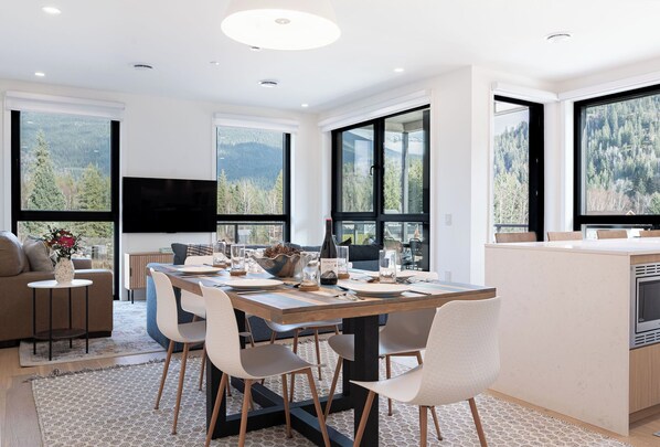 Un espace repas moderne avec une table en bois, des chaises blanches et une grande fenêtre donnant sur une vue imprenable sur les montagnes.