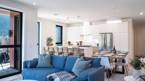 Un salon moderne avec un canapé bleu, un coin repas avec une table dressée pour quatre personnes, et une cuisine dotée de meubles blancs et d’appareils électroménagers en acier inoxydable.
