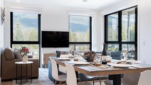 Un espace repas moderne avec une grande table dressée pour quatre personnes, entourée de chaises. Un téléviseur est fixé au mur au-dessus de la table.