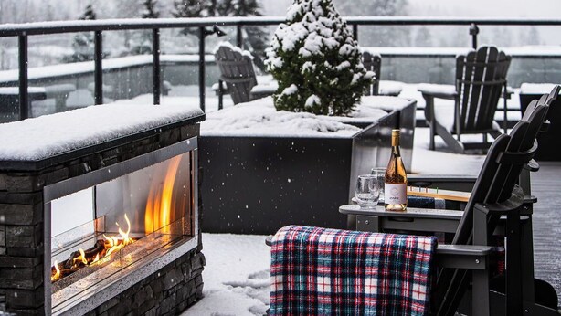 Une cheminée avec des flammes apparentes, un petit sapin de Noël, des sièges à l’extérieur et une terrasse enneigée.