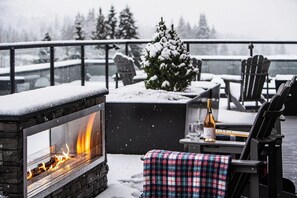 A fireplace with visible flames, a small Christmas tree, outdoor seating, and a snow-covered deck. - Wooley Bully Terrace  (Revelstoke)