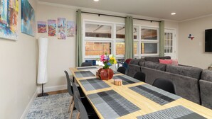 A dining area with a long table, chairs, and a vase with flowers. There is a couch and a television in the background. - 
Fenced Yard, Workspace, Cornhole + Life-Size Jenga (Arlington)