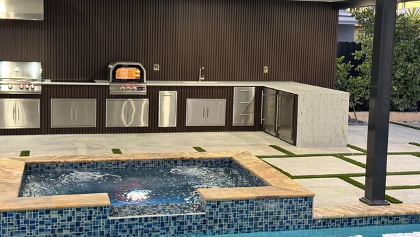 Et moderne udendørs køkken med indbygget grill, ovn og mikroovn, beliggende ved siden af en swimmingpool med fliseterrasse.
