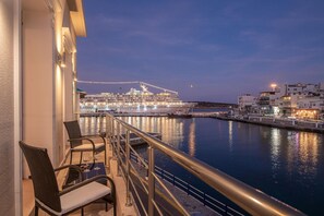 A balcony with wicker chairs and a table, overlooking a large cruise ship docked at a port with buildings in the background. - Sea View Petralis Residence   2 Bedroom apartment (Agios Nikolaos)