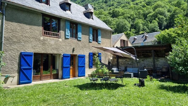 Une maison en pierre à deux étages, avec des volets bleus, une terrasse dotée de mobilier d’extérieur et un paysage montagneux en toile de fond.